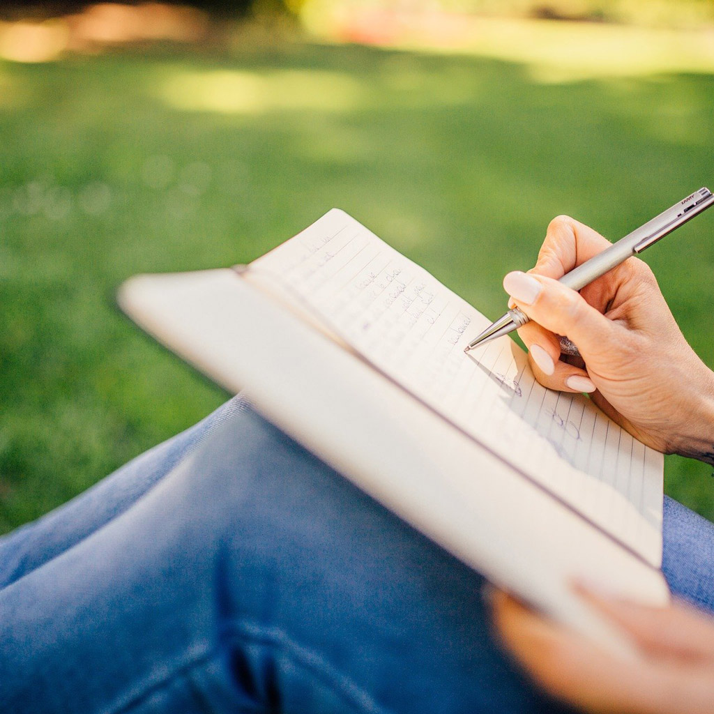 Close up of a woman sitting on grass writing in a notebook.