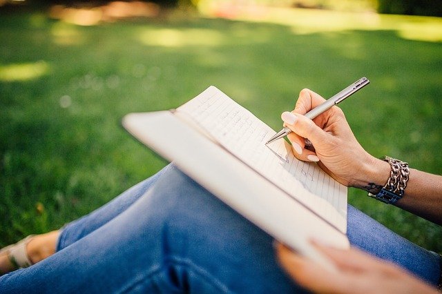 Close up of a woman sitting on grass writing in a notebook.